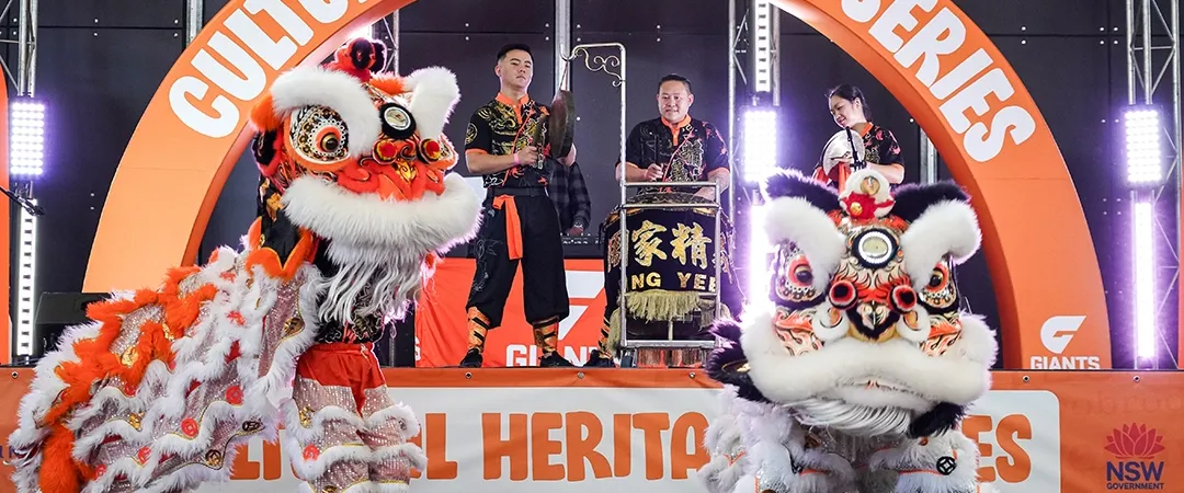 Lion dancers in vibrant orange and white costumes perform energetically on stage under the "Cultural Heritage Series" banner, with musicians playing behind them.