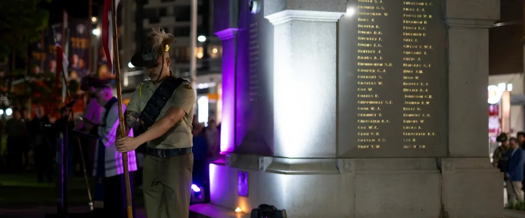 A person stands on a dimly lit street near a large stone monument, illuminated by purple and white lights, with an urban backdrop at night.