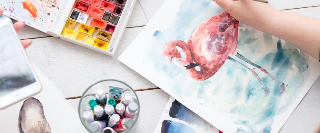 A hand paints a pink flamingo on white paper surrounded by watercolours and paint tubes on a table. The scene is vibrant and creative.