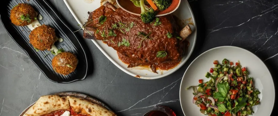 Grey table with plates of food like pizza, salad, roasted meat and vegetables. 