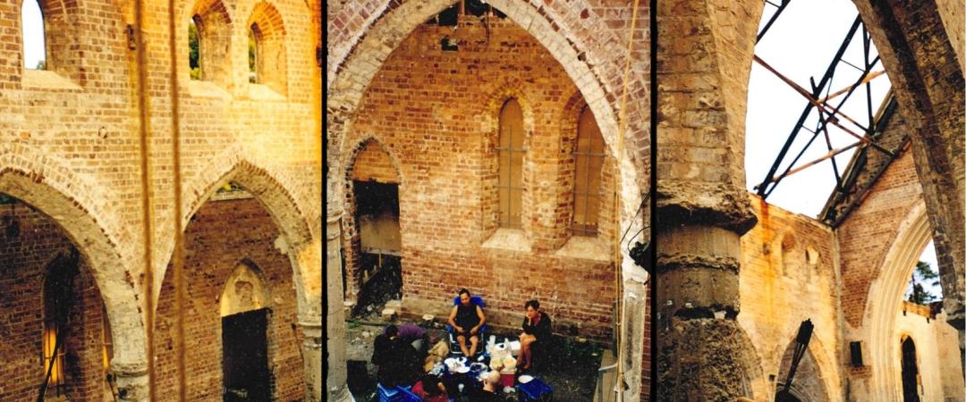 Group of people sit around a table in a roofless, abandoned brick church with arched windows. The atmosphere is relaxed and historic.