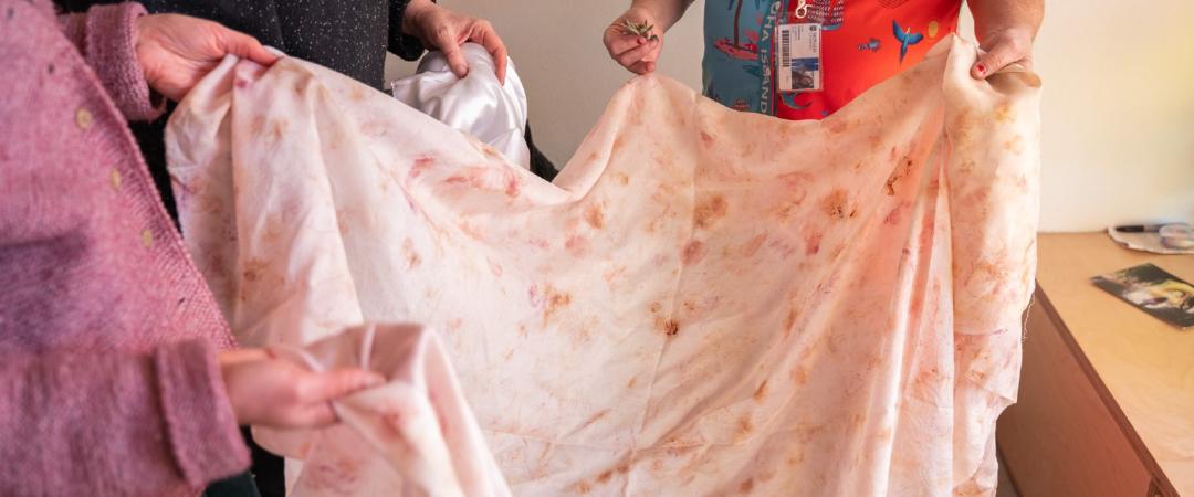 Three people hold up fabric with brownish, pink splotches on it from botanical dyes
