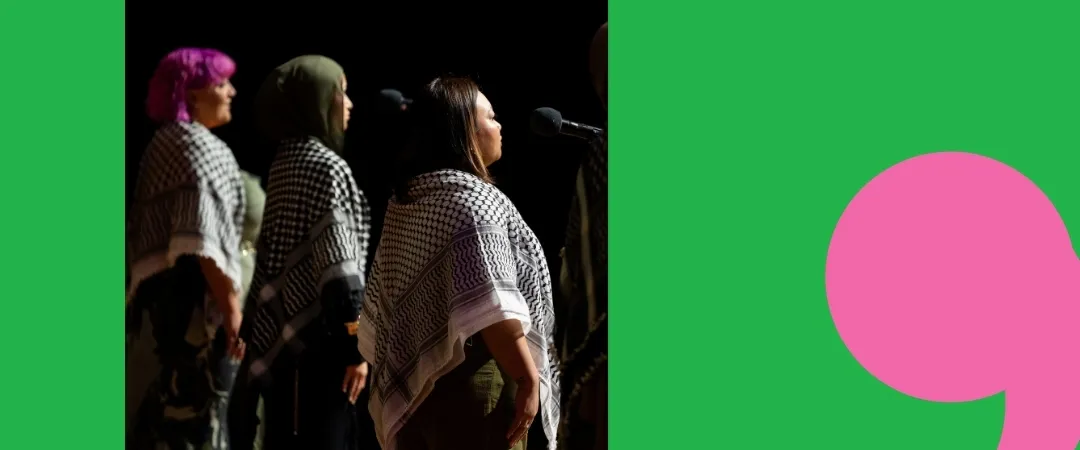 Four women stand side by side wearing black and white checkered shawls, facing microphones. The background is green with a large pink comma.