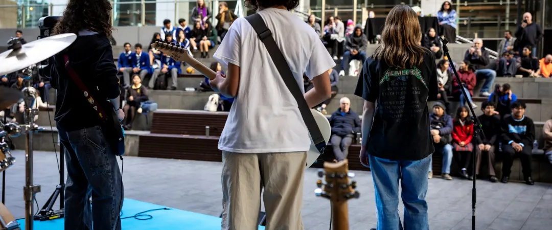 Three musicians perform on an outdoor stage, facing a large, seated audience. The lively atmosphere suggests enjoyment and engagement.