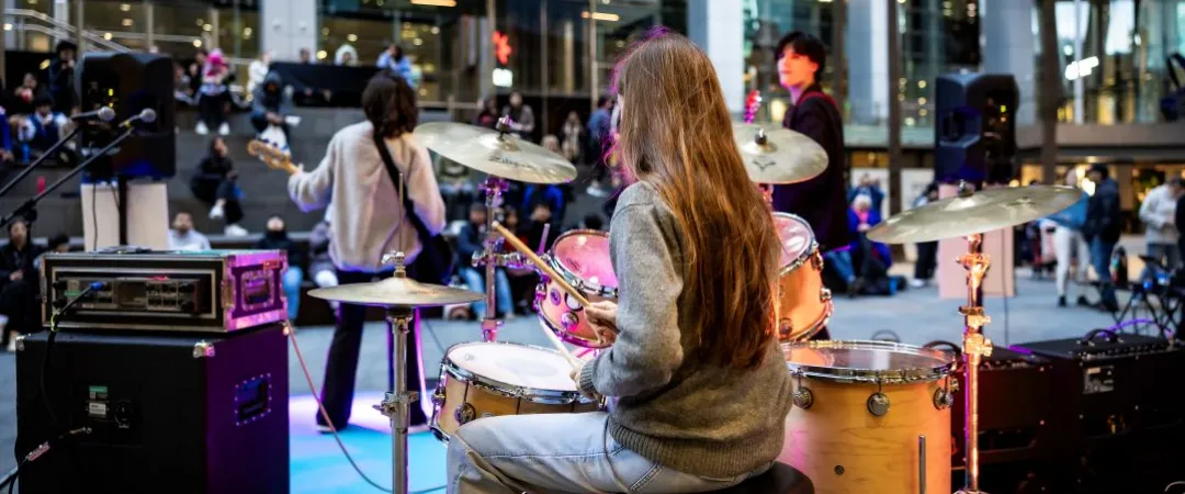 A street band performs in an urban setting. The drummer, in a grey sweater, plays energetically. Audience members sit on steps, watching the lively scene.