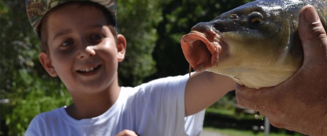 A young child smiles excitedly while holding a caught fish with help, outdoors on a sunny day. Lush greenery is visible in the blurred background.