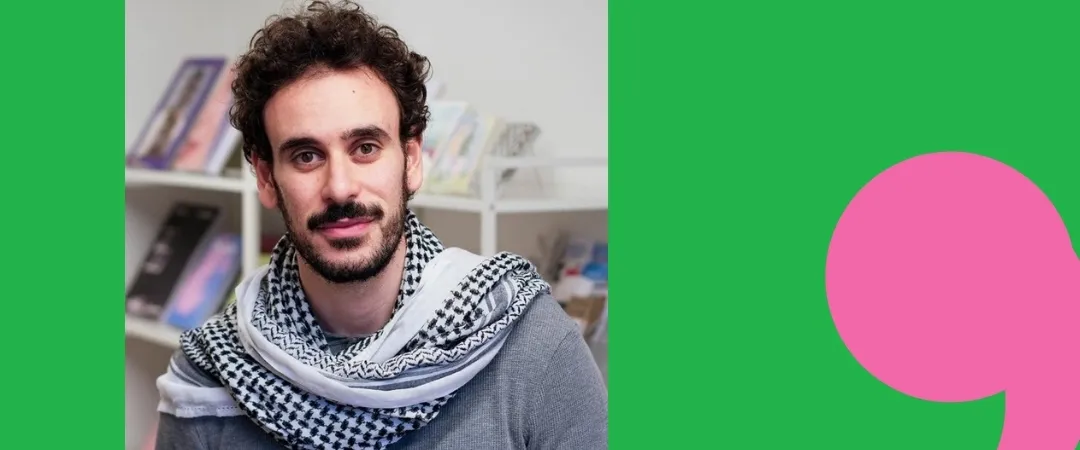 A man with curly hair and a beard wears a patterned scarf and gray sweater, standing in front of bookshelves. The background is green with a large pink comma.