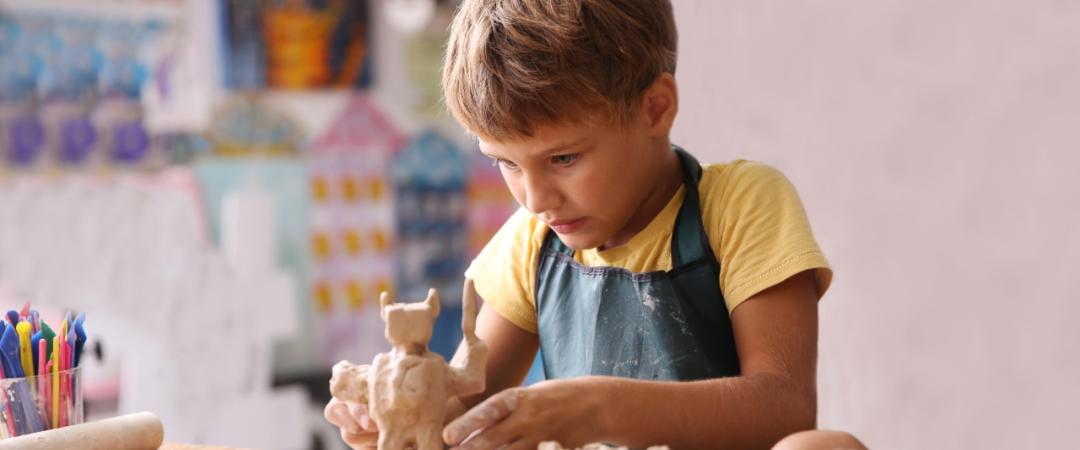 A focused child in a yellow shirt and apron sculpts clay at a table, surrounded by colourful art supplies, in a vibrant, creative studio setting.
