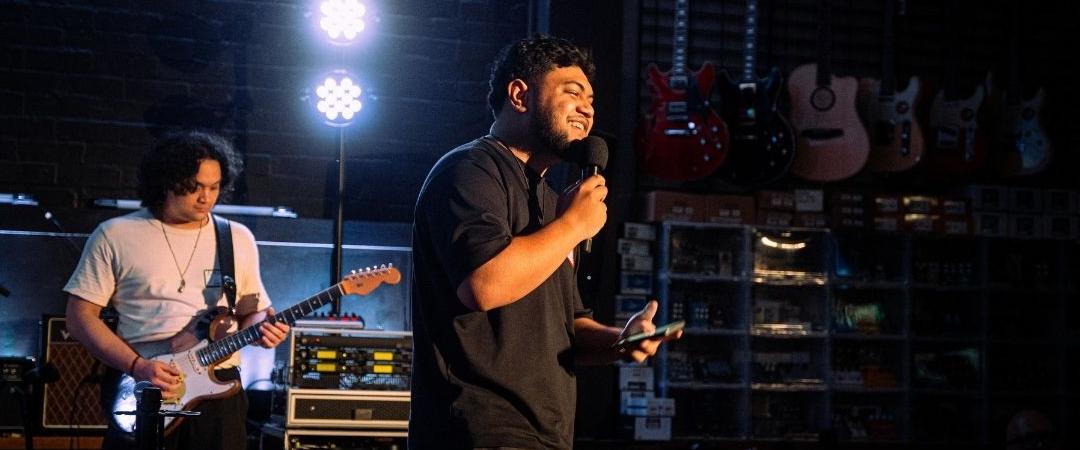 A rock band performs energetically on stage. The lead singer grips the mic, passionately singing, while the guitarist in a white shirt plays under bright stage lights.