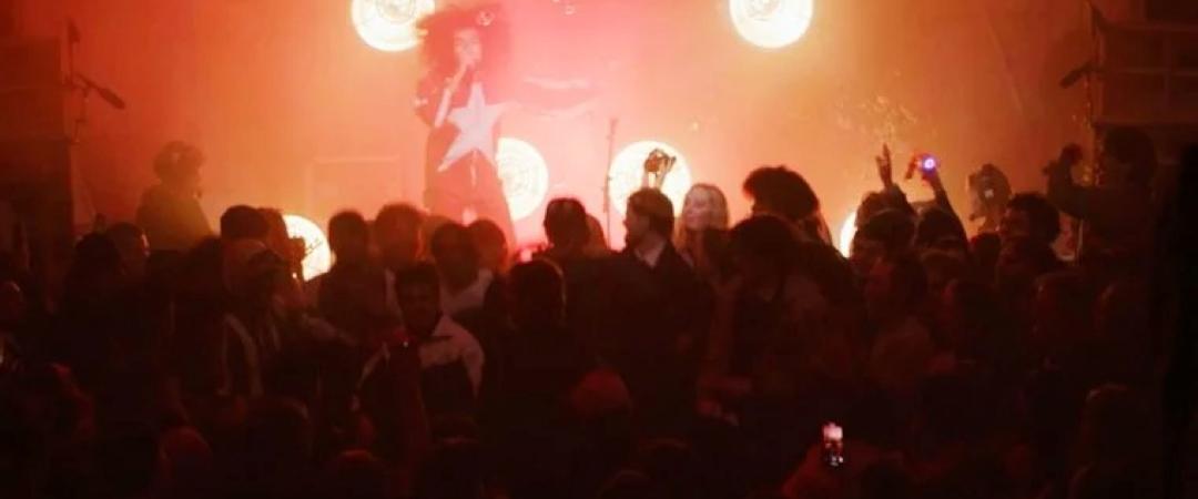 A performer stands on stage, backlit by intense red lighting and glowing circular lights. A large crowd is gathered, creating an energetic concert atmosphere.