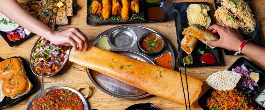 Overhead view of a vibrant Indian feast: long dosa, curries, rice, chutneys, and shared dishes on a wooden table, with hands serving food.