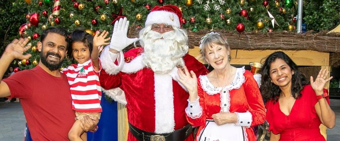 A family stands joyfully with Santa and Mrs. Claus in festive attire. Everyone is smiling and waving, conveying a warm, cheerful holiday spirit.