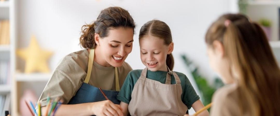 A mother and two daughters joyfully paint together at a table covered with art supplies. Colourful bunting decorates the background, creating a cheerful atmosphere.