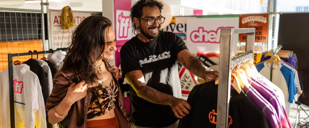 A couple smiles while browsing clothes in a cosy second-hand shop. Racks of colourful garments surround them, creating a cheerful and relaxed atmosphere.