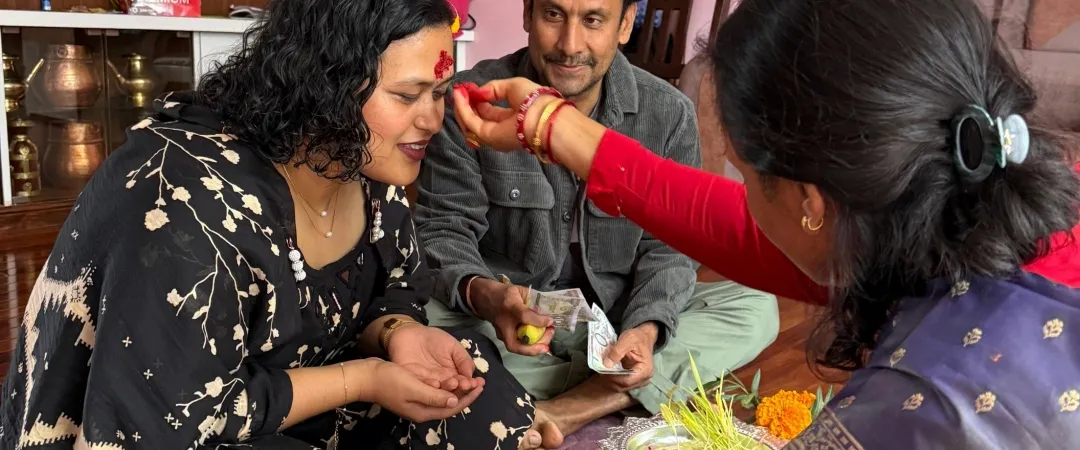 At a family ceremony, a woman in a purple sari applies a red tilak to a seated woman as a man watches; flowers and ritual offerings sit nearby.