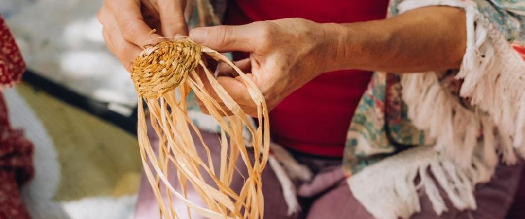 Hands of an elderly person weaving straw, wearing a colourful sweater. The scene conveys a sense of craftsmanship and tradition.