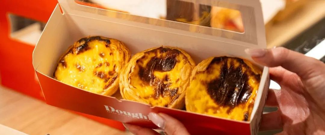 Hands hold a red bakery box containing three golden, caramelized egg tarts, warmly lit and ready to serve, creating an inviting mood.