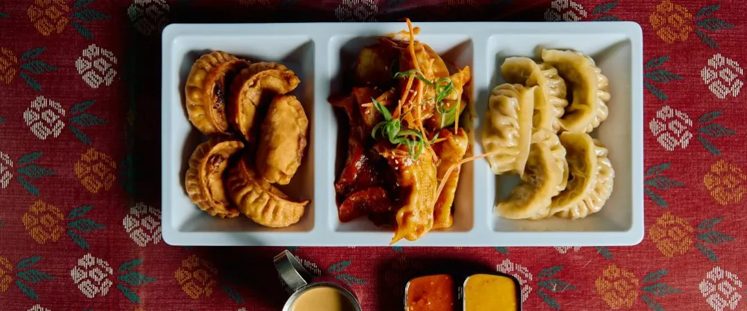 Overhead view of an Asian meal: fried pastries, shrimp in red sauce, steamed dumplings, and two dipping sauces on patterned red cloth.