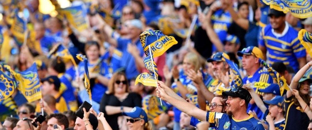 A crowd of enthusiastic sports fans in blue and yellow jerseys cheer and wave flags passionately, creating a lively and energetic atmosphere.