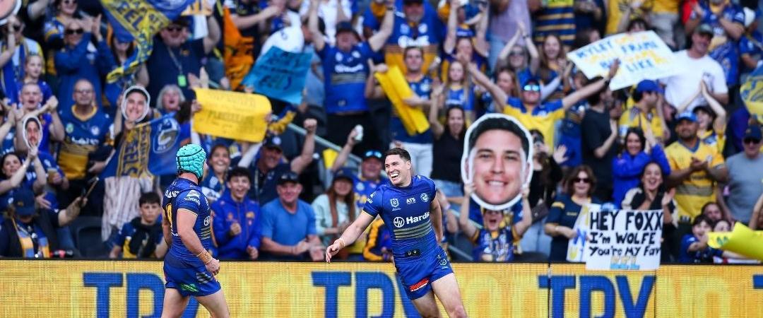 Two football players in blue jerseys run on a field with a cheering crowd. Fans hold signs and banners, creating an energetic and celebratory atmosphere.