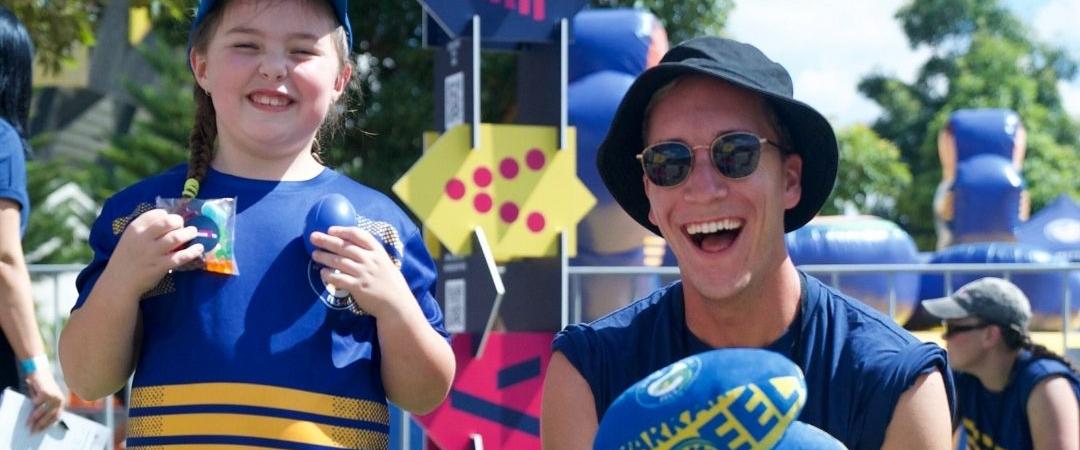 Two people smiling at a street festival, wearing blue and yellow outfits and bucket hats. Colourful lanterns and trees are in the background.