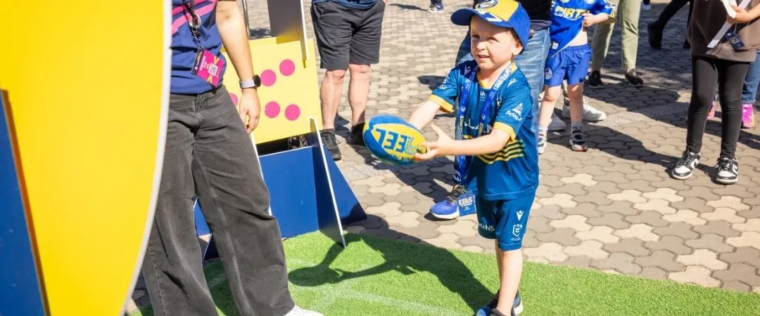 A joyful child in a blue shirt and shorts throws a football, aiming at a target at an outdoor event. Attendees watch, and the mood is lively and playful.