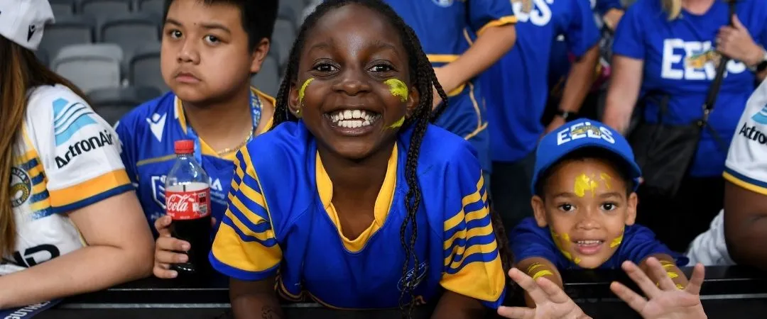 Excited children in blue sports jerseys lean against a stadium barrier, smiling and cheering. The atmosphere is lively and energetic.