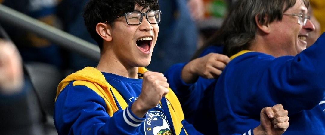 A young fan in a blue and yellow outfit with glasses cheers enthusiastically at a sports event, surrounded by other excited people. The atmosphere is lively.