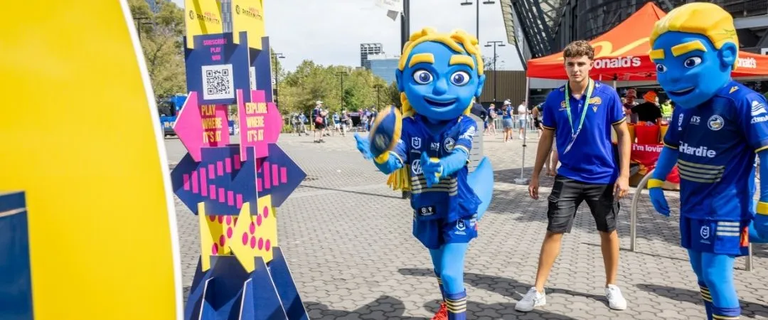 Two people in vibrant blue dinosaur costumes stand near a colourful game display outdoors. Nearby, someone in casual clothing appears cheerful.
