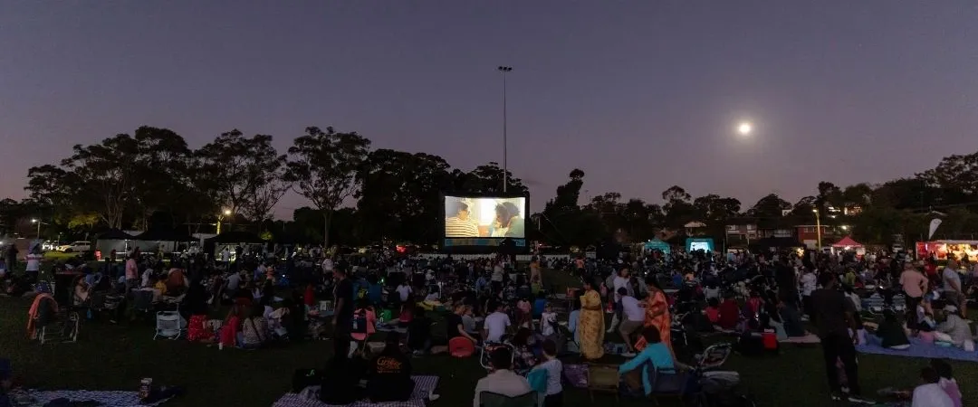 Outdoor movie night in a park under a clear sky. A crowd on blankets and lawn chairs watches a screen. The moon glows brightly, creating a serene atmosphere.
