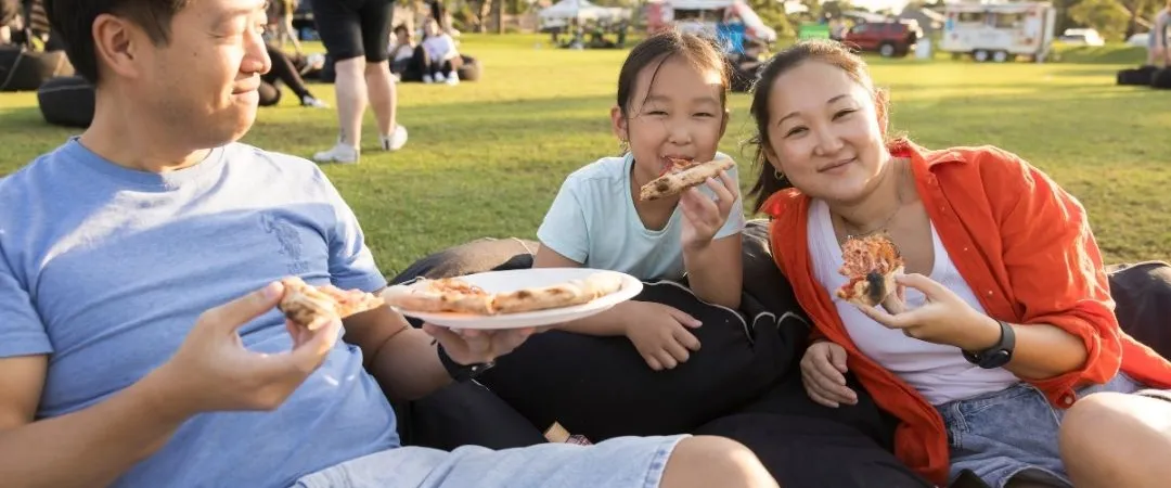 A family of three enjoys a picnic outdoors, smiling and eating sandwiches. They sit on a blanket in a sunny park, creating a joyful and relaxed atmosphere.