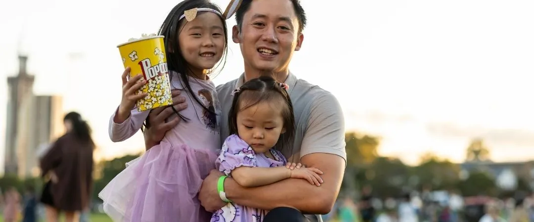 A smiling adult holds two children wearing purple dresses in a grassy park. The children hold popcorn and look happy; people and buildings are in the background.