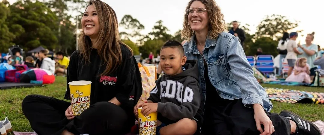 Three people sit on a pink blanket at an outdoor event. They're smiling and holding snacks, with trees and a crowd in the background, conveying a joyful mood.