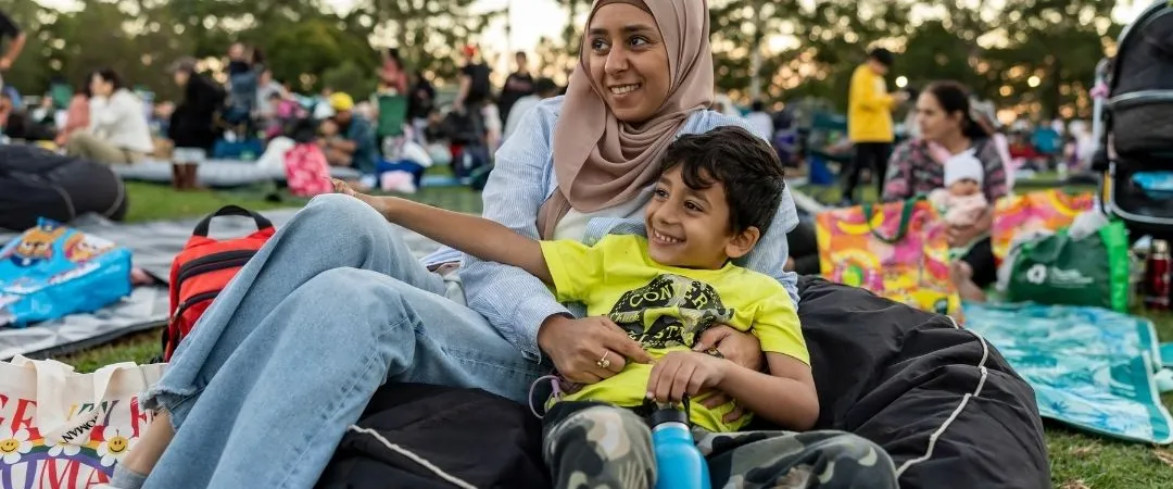 A woman and child sit on a blanket in a park, surrounded by people enjoying a picnic. The woman smiles warmly, creating a relaxed, joyful atmosphere.