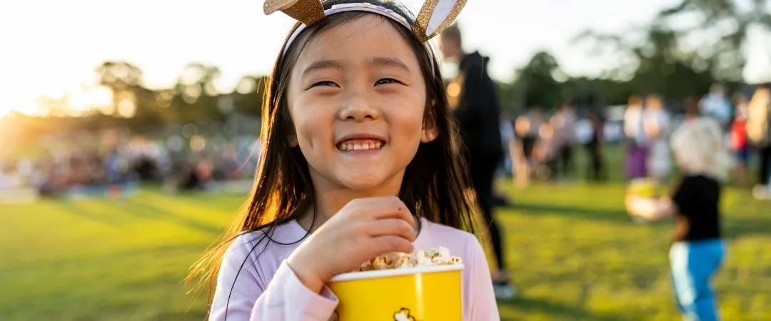 A smiling girl wearing cat ears enjoys popcorn at an outdoor event. Sunlight casts a warm glow as people mingle on the grass in the background.