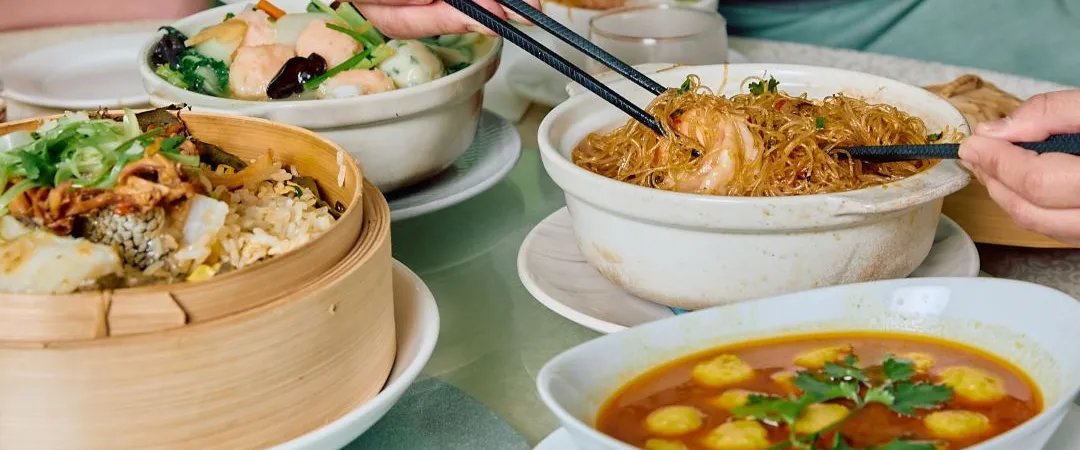 Diners share a Chinese meal, using chopsticks and spoons to serve noodles, dumplings, rice, vegetables, and soup at a bustling table.