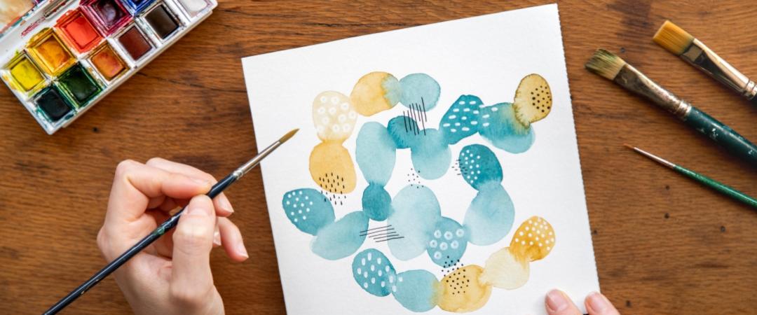Hand painting abstract watercolor circles in teal and yellow on white paper. Nearby are a watercolour palette and brushes on a wooden table.
