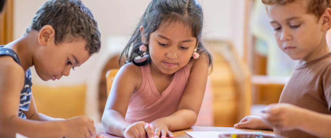 Three children are sitting around a table, drawing with crayons on paper. They appear focused and engaged, sharing colors from a basket.