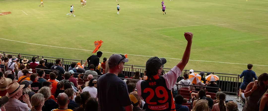 A lively rugby match with a cheering crowd. Fans in orange and black fill the stands. A person, wearing number 29, raises a fist in excitement as players engage on the field.