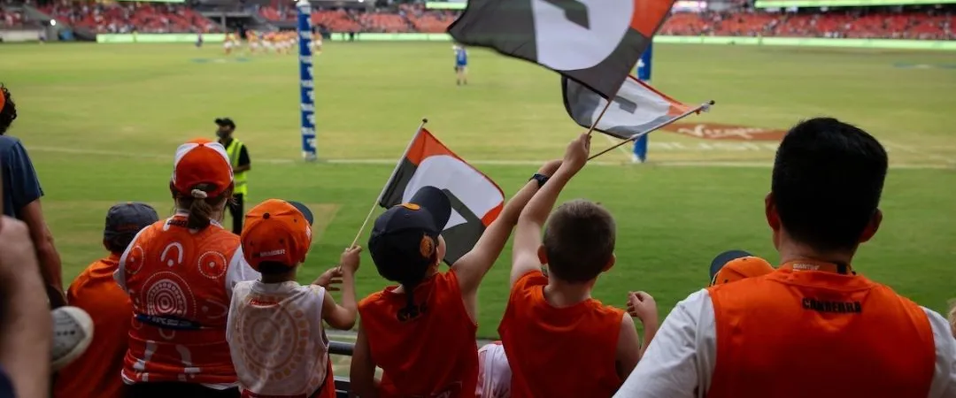 Fans dressed in red cheer enthusiastically, waving flags in a stadium. The atmosphere is lively, with anticipation in the air for the sporting event.