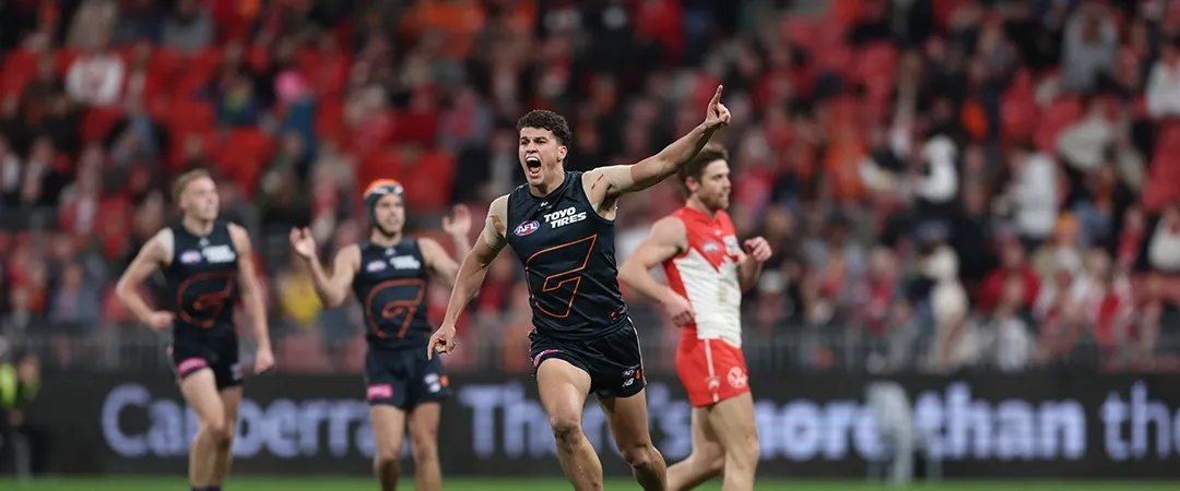 A jubilant Australian Rules football player in a dark jersey, number 7, celebrates a goal with arm raised on a field, surrounded by cheering teammates and an opponent in red.
