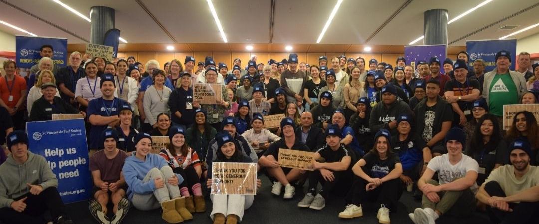 Large group of people in round sitting down & smiling for the camera all wearing blue beanies and signs like "Help us help people in need"