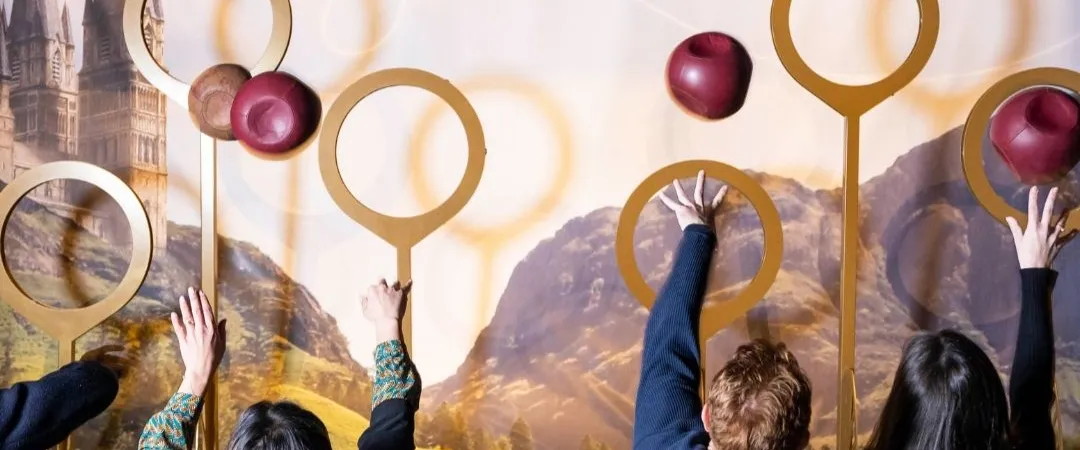 Two people wearing robes reach up to grab red balls in front of a mural depicting hoops. The scene evokes excitement and whimsy from a fantasy world.