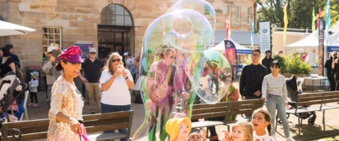 A street performer creates large bubbles in a lively outdoor market. People watch, smiling, near stalls with colorful items. A sunny, cheerful day.