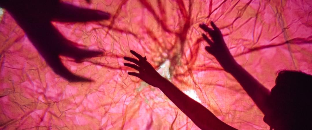 Silhouetted hands reach towards each other against a vibrant pink backdrop, creating a dramatic, mysterious atmosphere with strong shadow play.
