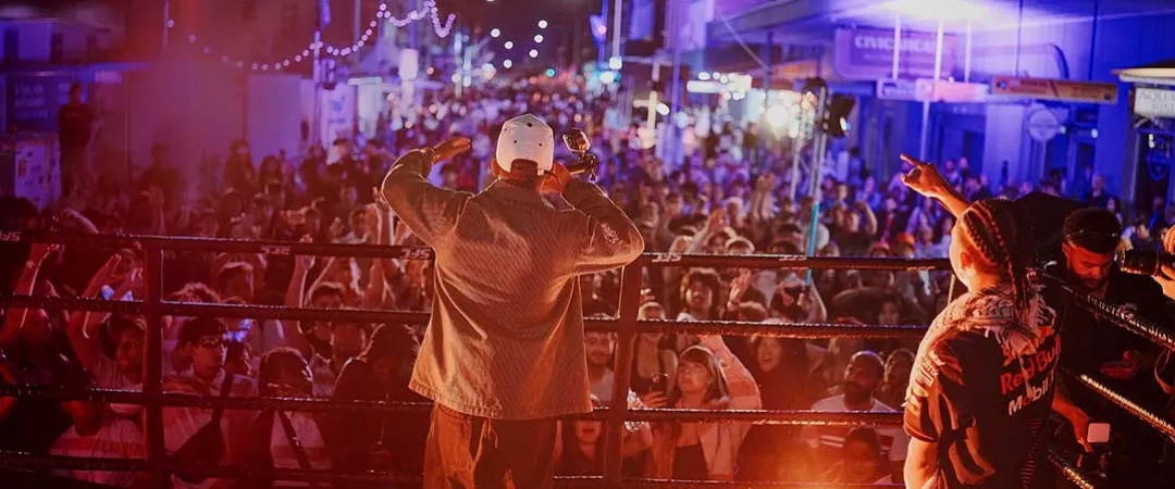 A performer in a white cap faces a cheering crowd from a stage at night, joined by dancers under purple and orange concert lights.