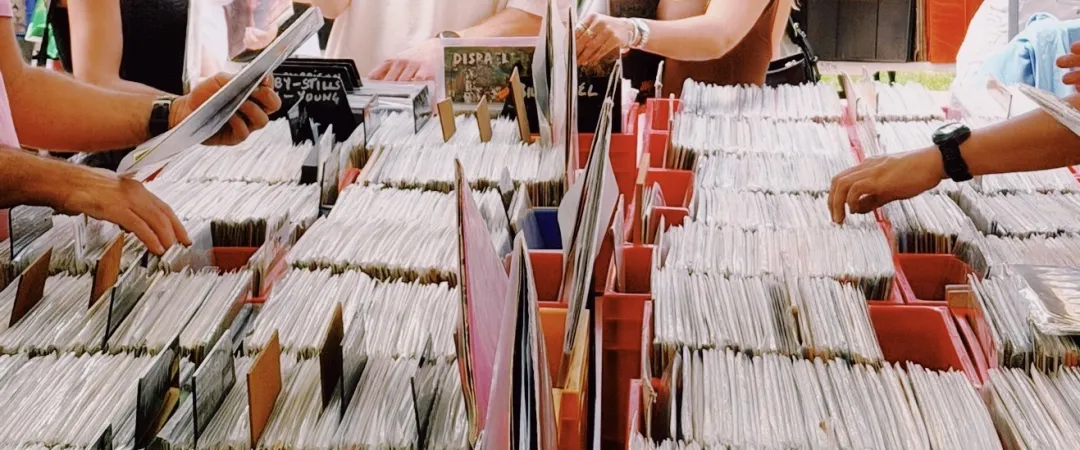 Hands browse through rows of vinyl records in colorful bins at a busy market. The scene conveys a nostalgic and lively atmosphere.
