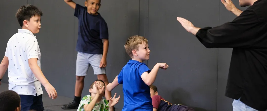 Children playfully follow an instructor’s gestures in a classroom, with some standing and others seated in a lively, joyful group activity.