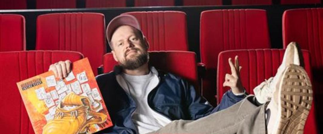 A person lounges casually in red theater seats, holding a vinyl record. They appear relaxed and joyful, with feet propped up and a big smile.