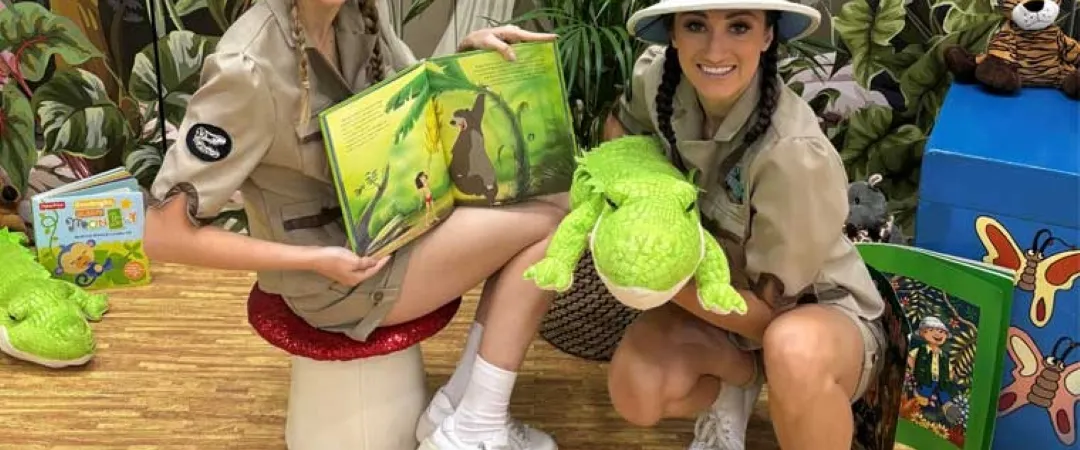 Two smiling women in safari hats pose with a jungle-themed book and plush crocodiles amid lush plants, creating a playful, educational scene.